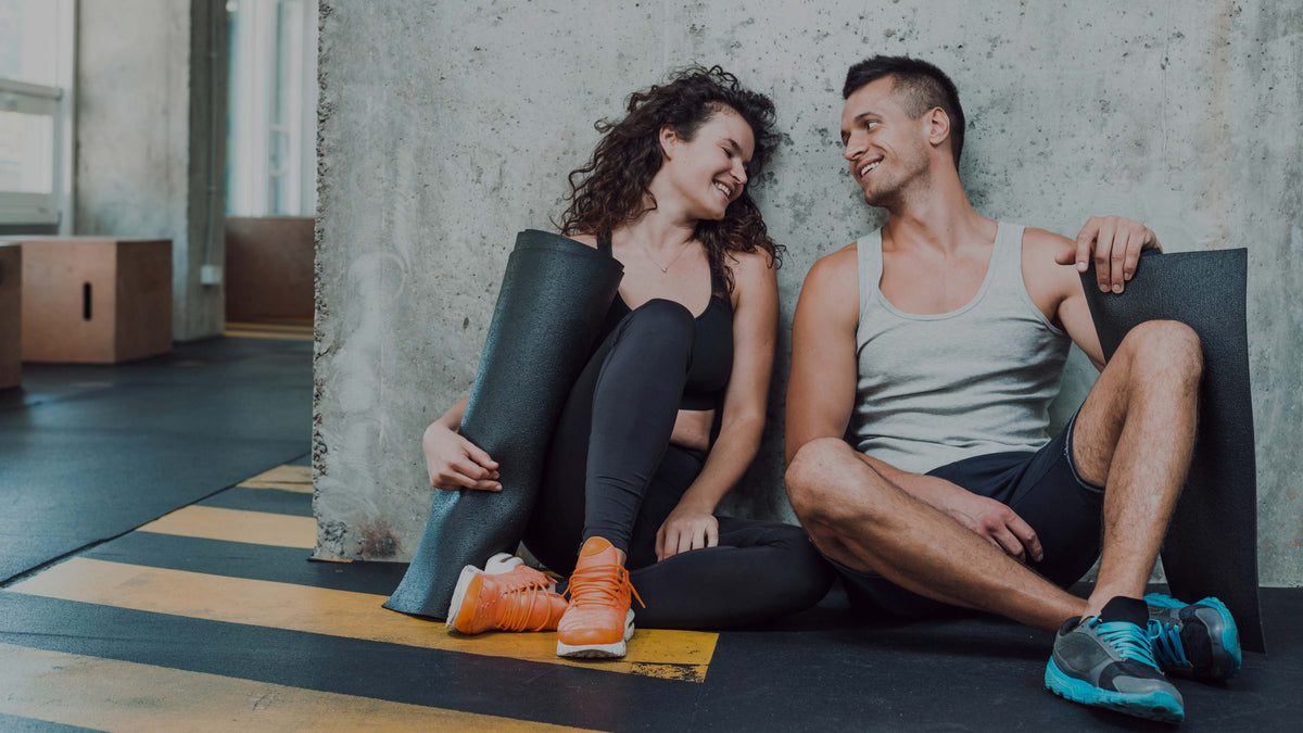 A smiling couple sits on a gym floor against a concrete wall, each holding a black exercise mat. The woman wears a black sports bra and leggings with bright orange sneakers, and the man wears a light gray tank top, dark shorts, and blue sneakers.