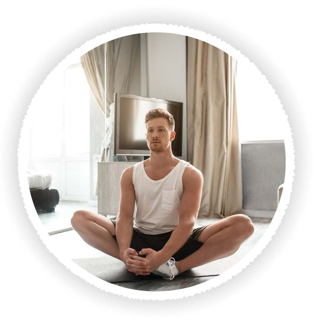 A man in a white tank top and black shorts sits cross-legged on a yoga mat in a bright, modern living room setting, framed within a circular border on a white background.