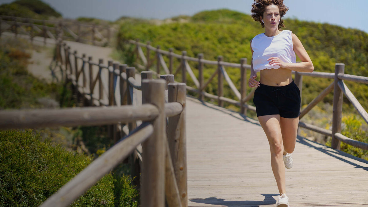 A woman jogging on a wooden boardwalk path outdoors, wearing a white sleeveless top, black athletic shorts, and white sneakers.