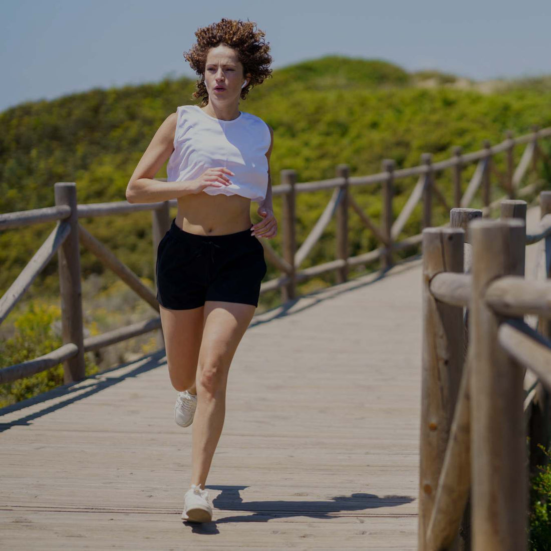 A woman jogging on a wooden boardwalk outdoors, wearing a white sleeveless crop top, black athletic shorts, and white sneakers.