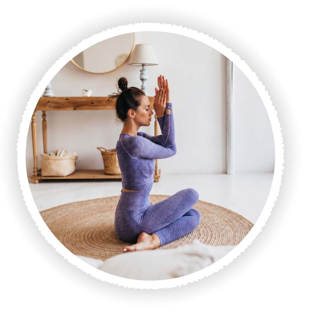 A woman in a matching purple yoga set sits cross-legged on a woven rug in a bright room, holding her hands together in a prayer pose. The photo is framed within a white scalloped circular border.